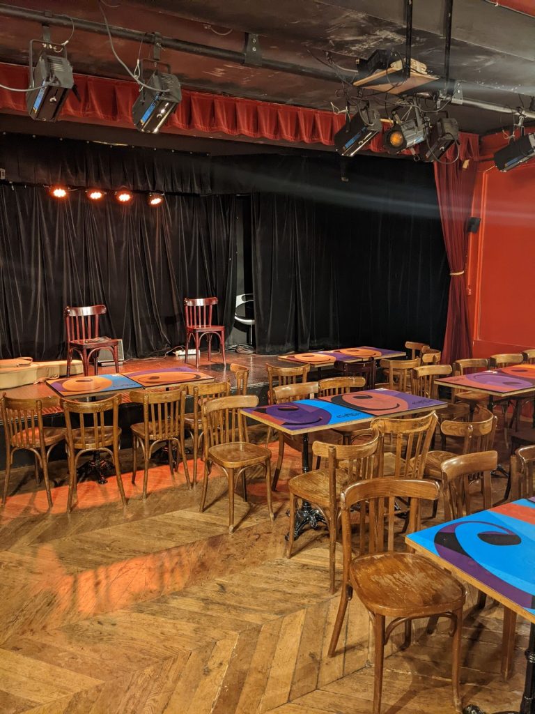 Salle de spectacle du Complexe du Rire avant l’arrivée des invités : scène aux rideaux noirs, tables bistrot colorées et chaises en bois