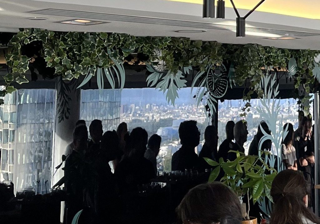 Invités en silhouette devant les baies vitrées et la vue sur la ville, sous une guirlande de lierre, au Skyline Bar du Meliá Paris La Défense.
