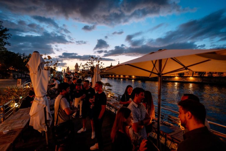 Invités de la soirée de lancement de WalletConnect sur la terrasse de la péniche La Balle au Bond à la nuit tombée, quais de Seine éclairés en arrière-plan, à Paris