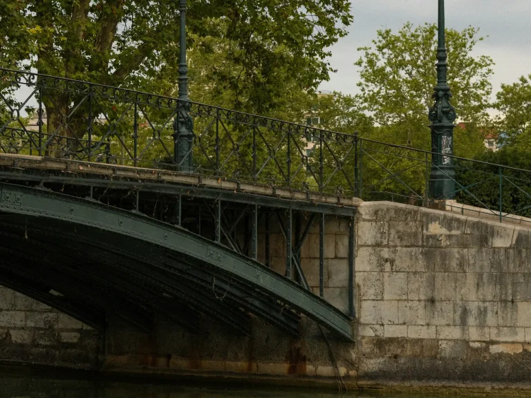 Un pont de fer et ses réverbères au-dessus de l’eau, sous les platanes, à Annecy