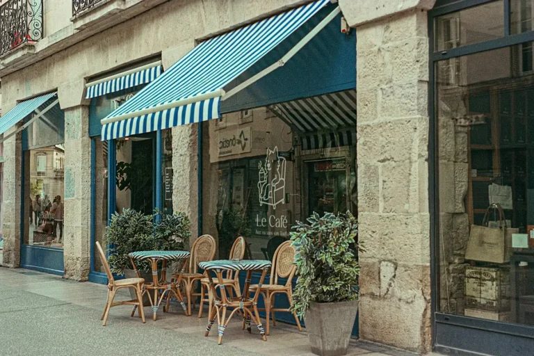 Tables et chaises d’une terrasse de café sous des stores rayés, devant une façade en pierre du centre de Grenoble