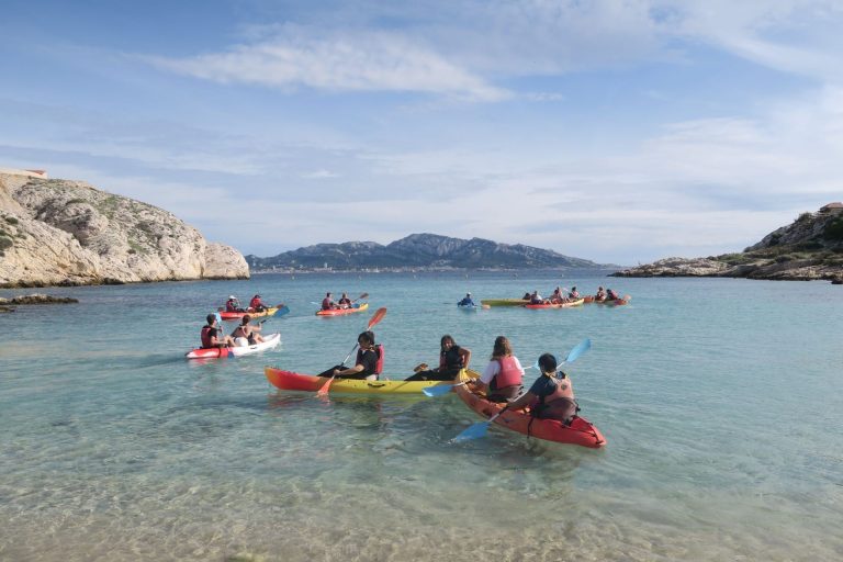 Swiss Life employees in two-seater kayaks in a clear-water cove of the Frioul islands, with the hills of Marseille in the distance, during their team building