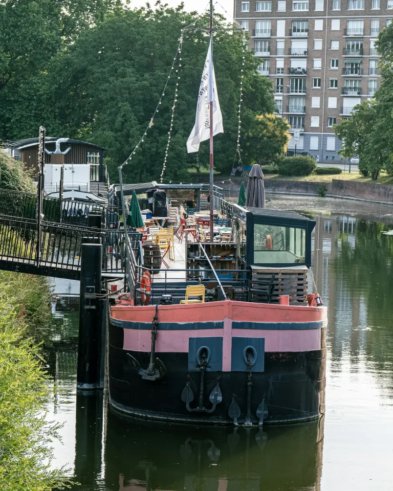 Une péniche amarrée au bord de la Deûle, à Lille, entre les arbres et une passerelle