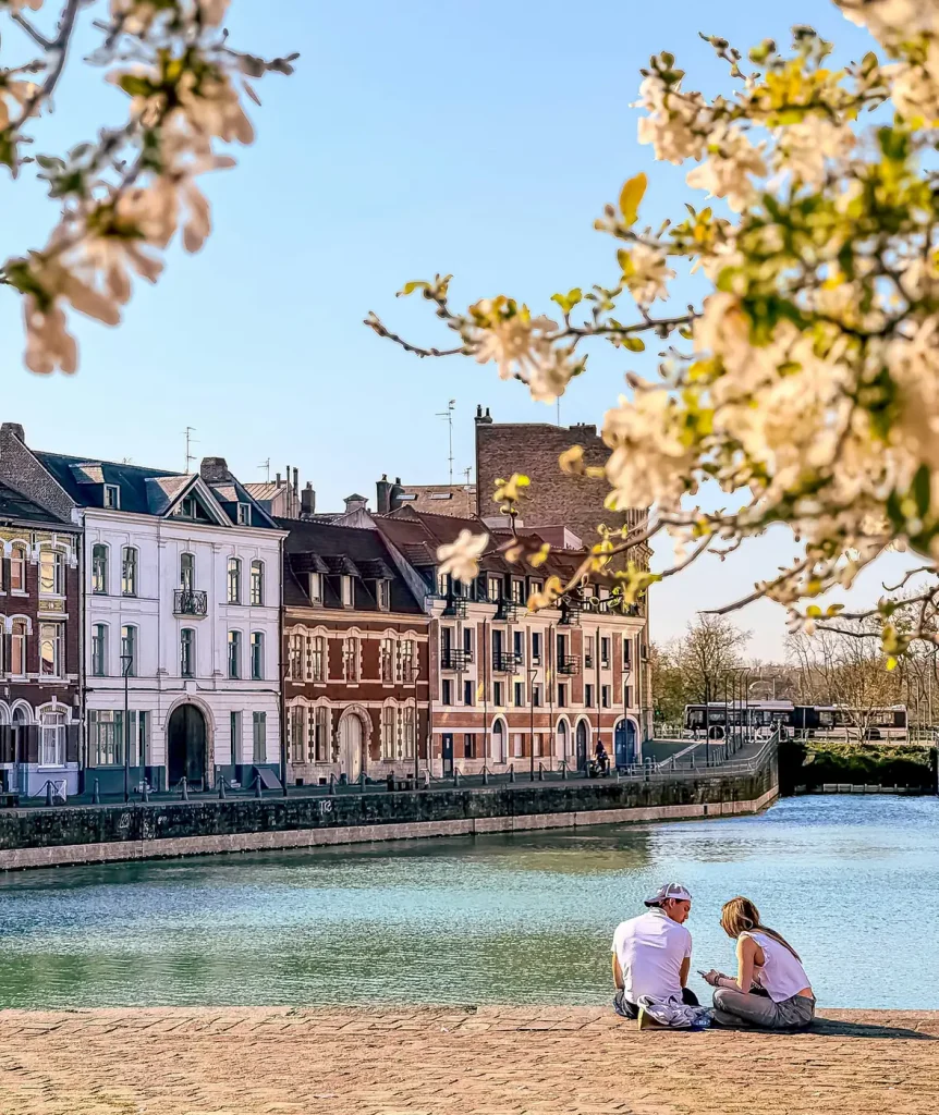Promeneurs assis au bord de l’eau, quai du Wault, à Lille, face aux façades colorées et sous des branches en fleurs