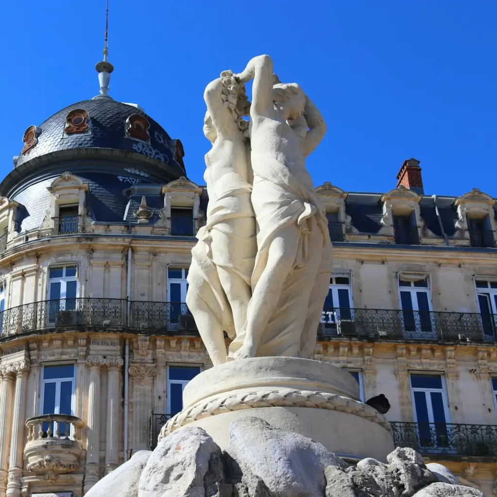 La fontaine des Trois Grâces sur la place de la Comédie, à Montpellier, devant une façade à dôme et balcons en fer forgé, sous un ciel bleu