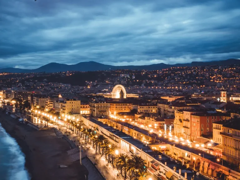 La Promenade des Anglais et le front de mer de Nice illuminés à la tombée de la nuit, avec les collines sous un ciel nuageux
