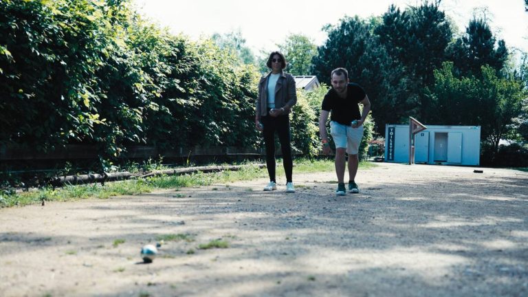 Deux participants du séminaire KPMG jouent à la pétanque sur une allée de gravier, entre les arbres du Center Parcs du Lac d’Ailette