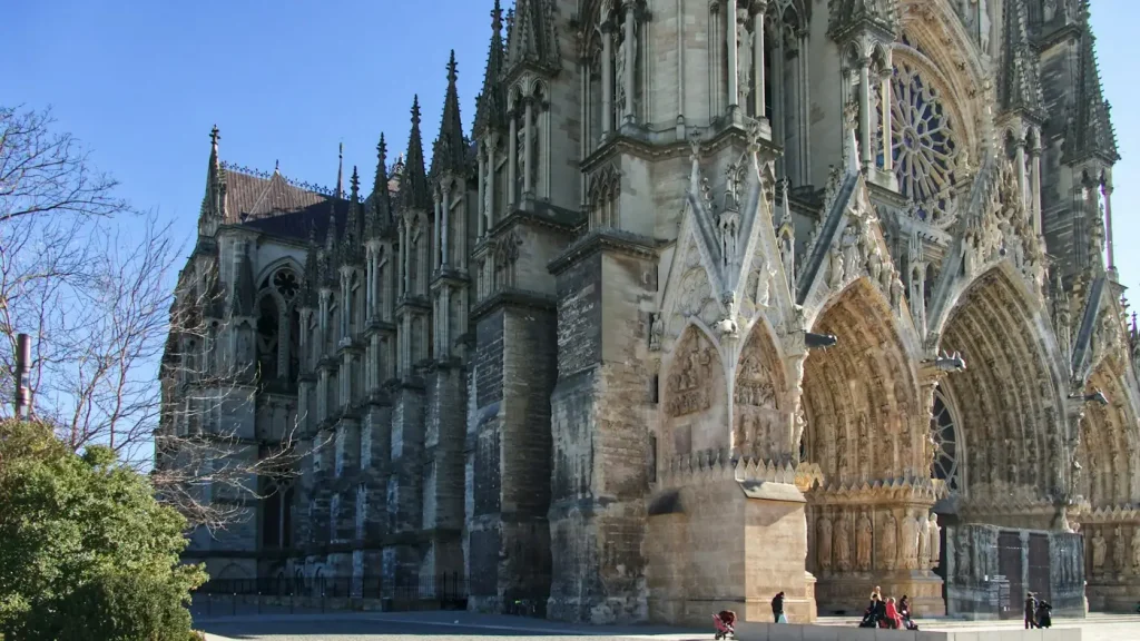 Le flanc et les portails sculptés de la cathédrale Notre-Dame de Reims, quelques passants sur le parvis, sous un ciel bleu