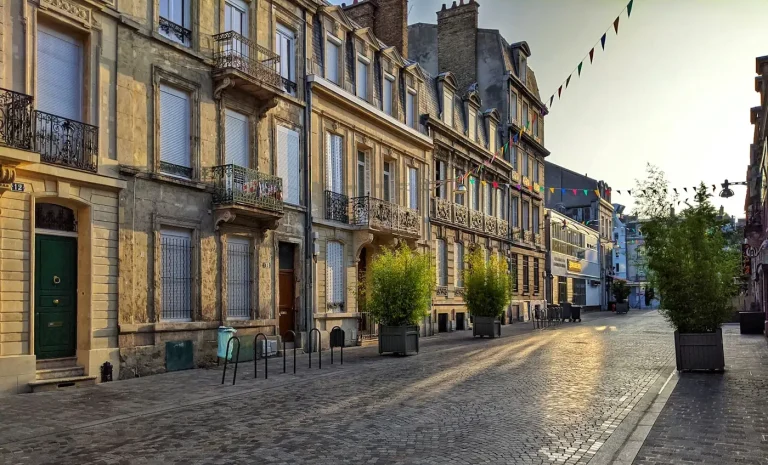 Rue pavée du centre de Reims, façades en pierre, bacs de plantes et fanions, au soleil du matin
