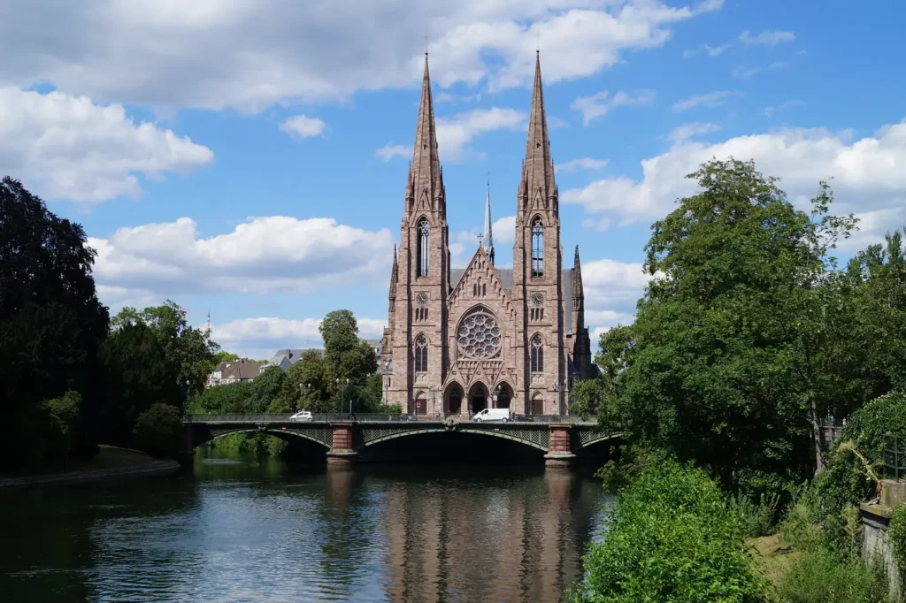 L’église Saint-Paul et ses deux flèches au bord de l’Ill, avec un pont de pierre, à Strasbourg, sous un ciel bleu