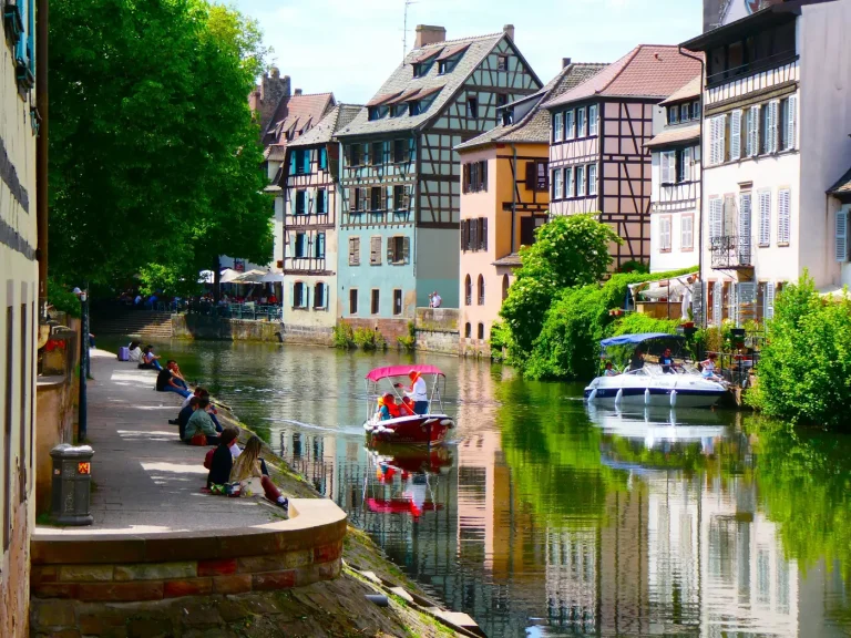 Promeneurs assis sur un quai de l’Ill et petits bateaux devant les maisons à colombages de la Petite France, à Strasbourg