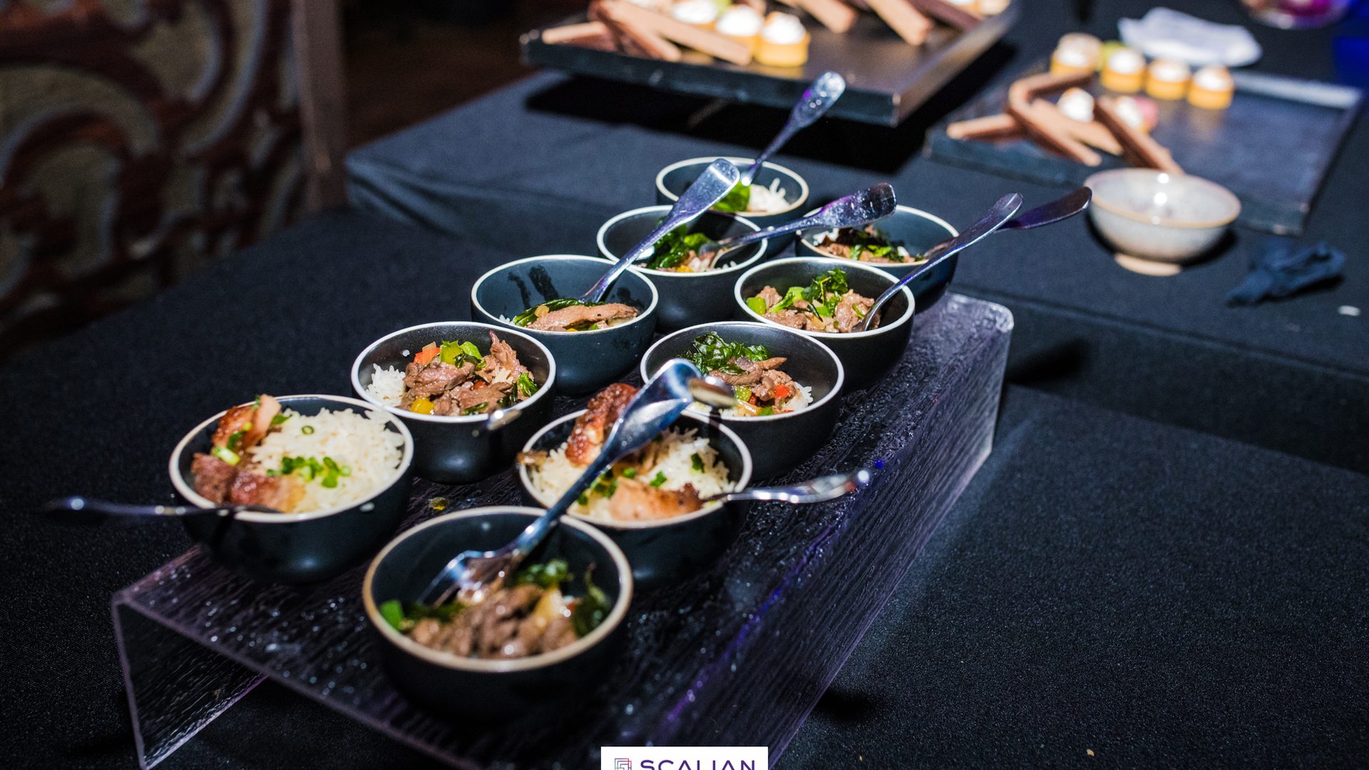 Bols de riz garnis de viande et d'herbes fraîches, présentés sur un présentoir transparent avec leurs cuillères, lors d'une soirée Scalian organisée par Weevup